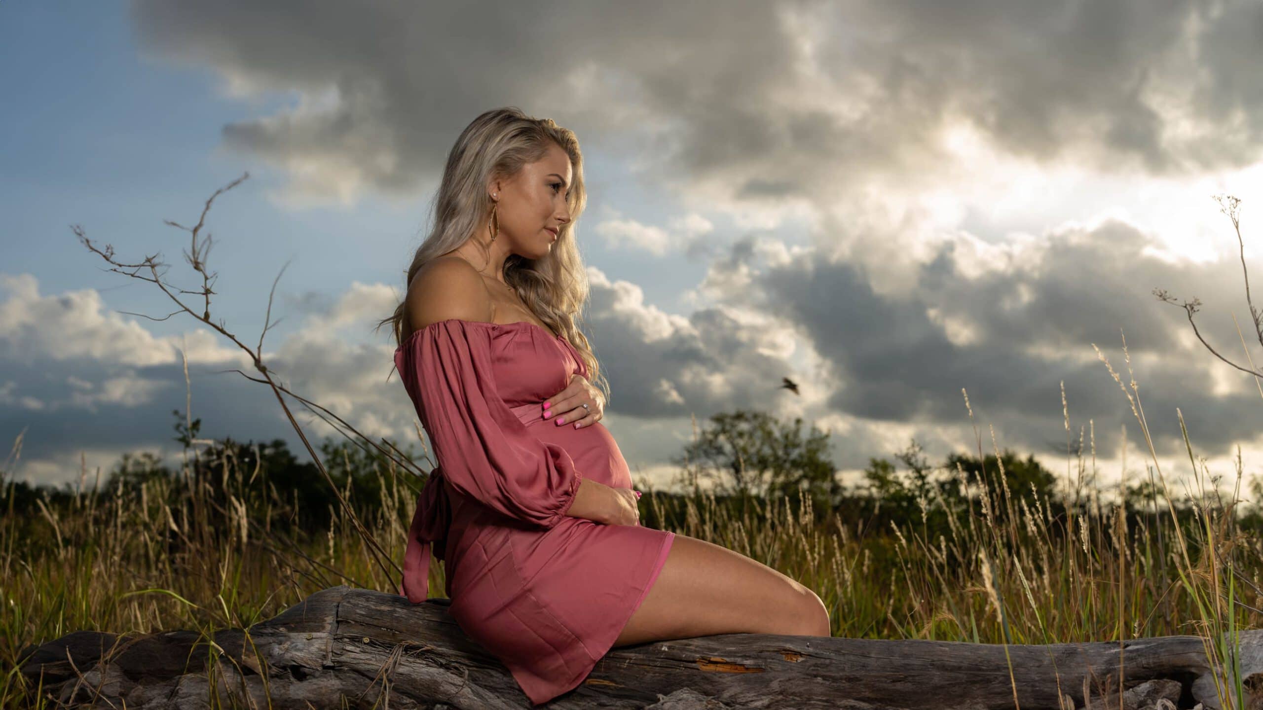 Pregnant woman sitting on log in field.