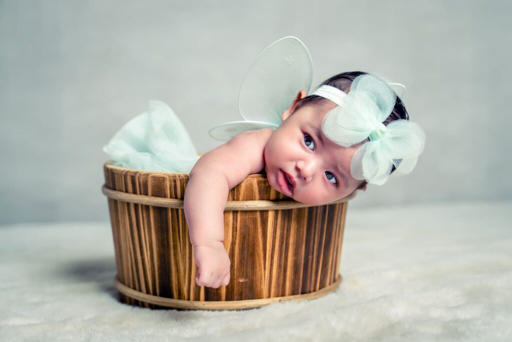Baby in wooden bucket wearing a bow in hair and fairy wings.