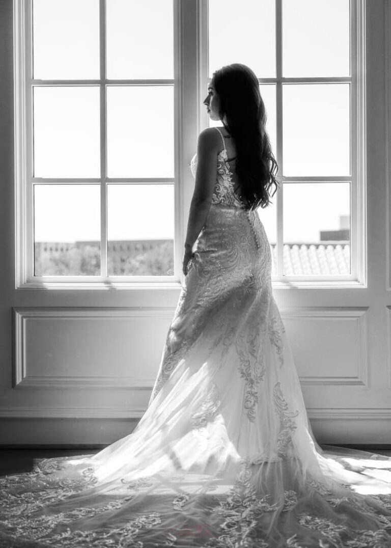 Bride poses for photo in dress by window.