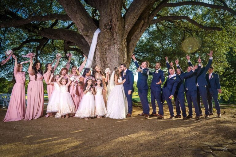 Wedding party celebrates and newlyweds kiss under tree.