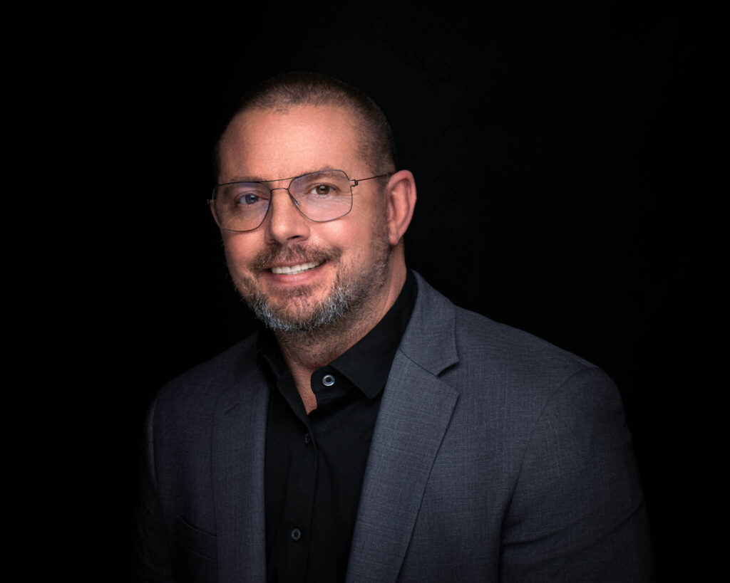 Headshot of a man in a great suit, black shirt and no tie and wearing glasses.