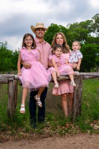 A couple and 3 kids wearing pink. Posing for a photo on an old wood fence.