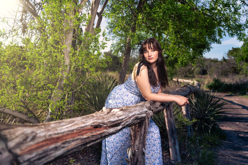 Senior portrait of a pretty girl with dark hair and blue eyes. Leaning on a fence with foliage in background.