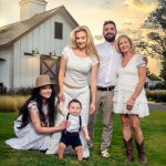 Grandmother, kids and grand baby pose in front of white barn at sunset.