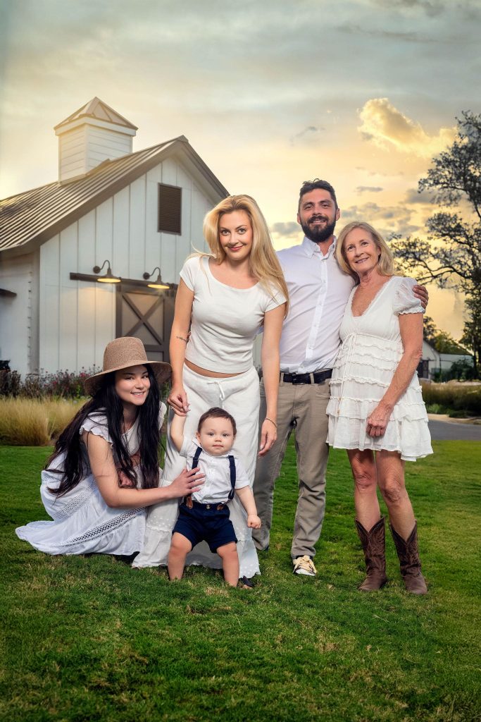 Grandmother, kids and grand baby pose in front of white barn at sunset.